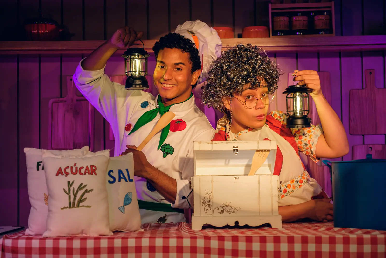 Jean Marques, como João, e Ana Pereira, como Vó Alzira, segurando lanternas atrás de um baú, em cena.